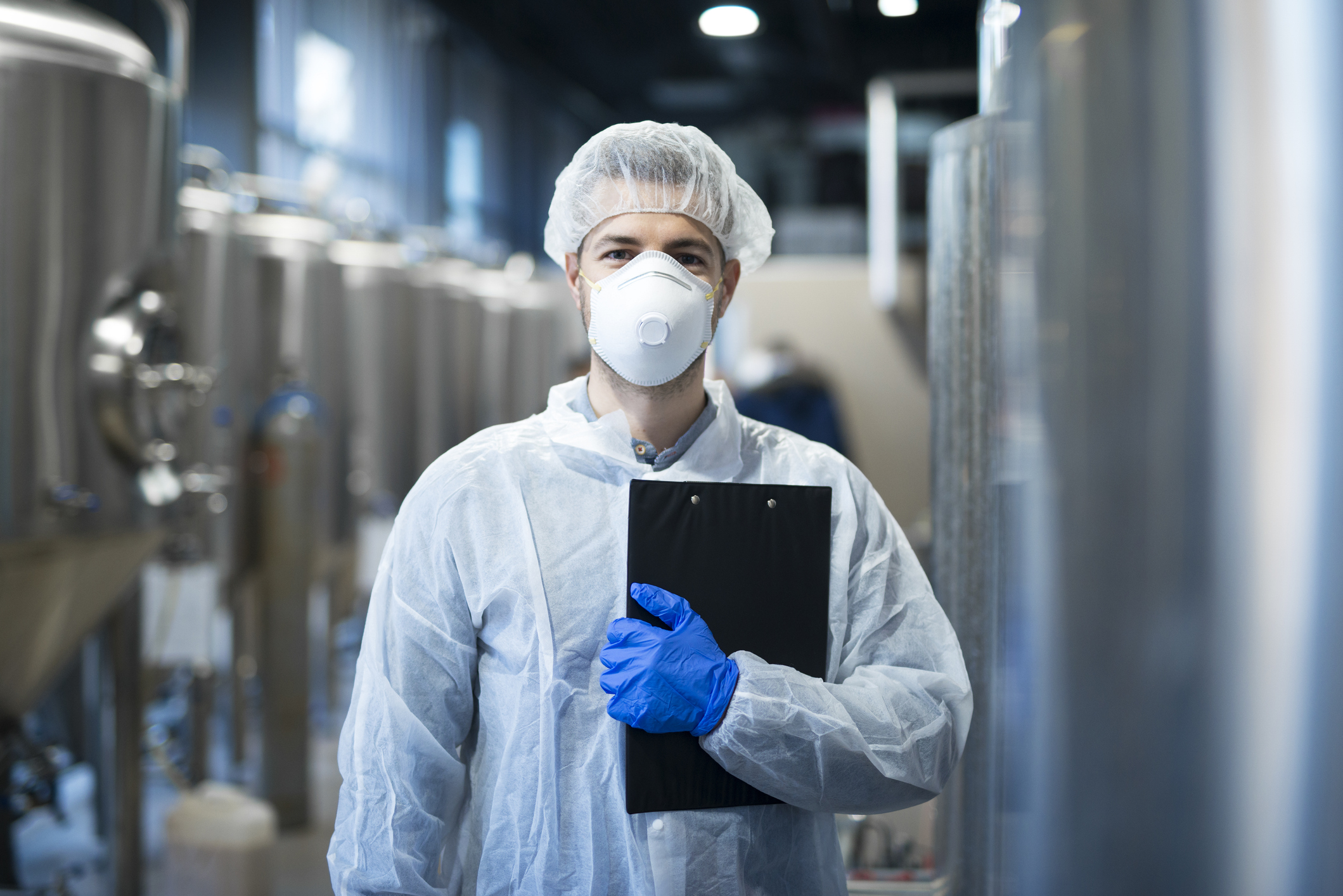 Technologist with protective mask and hairnet standing at factory production line. Food or beverage processing factory.